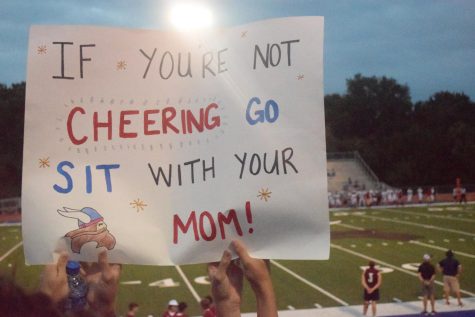 Senior Kynlea Heydenreich holds up a sign with her friend, stating, If youre not cheering go sit with your mom! Heydenreich has been a consistent participant in the Vikings student section during football, preparing signs and dressing up for all the themes.